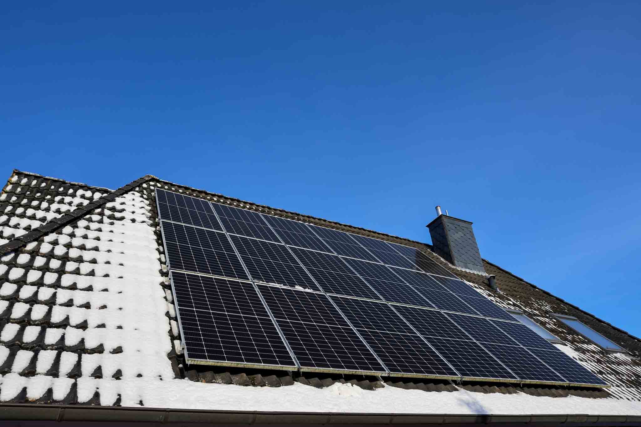 Solar panels on a house roof partially covered with snow against a clear blue sky