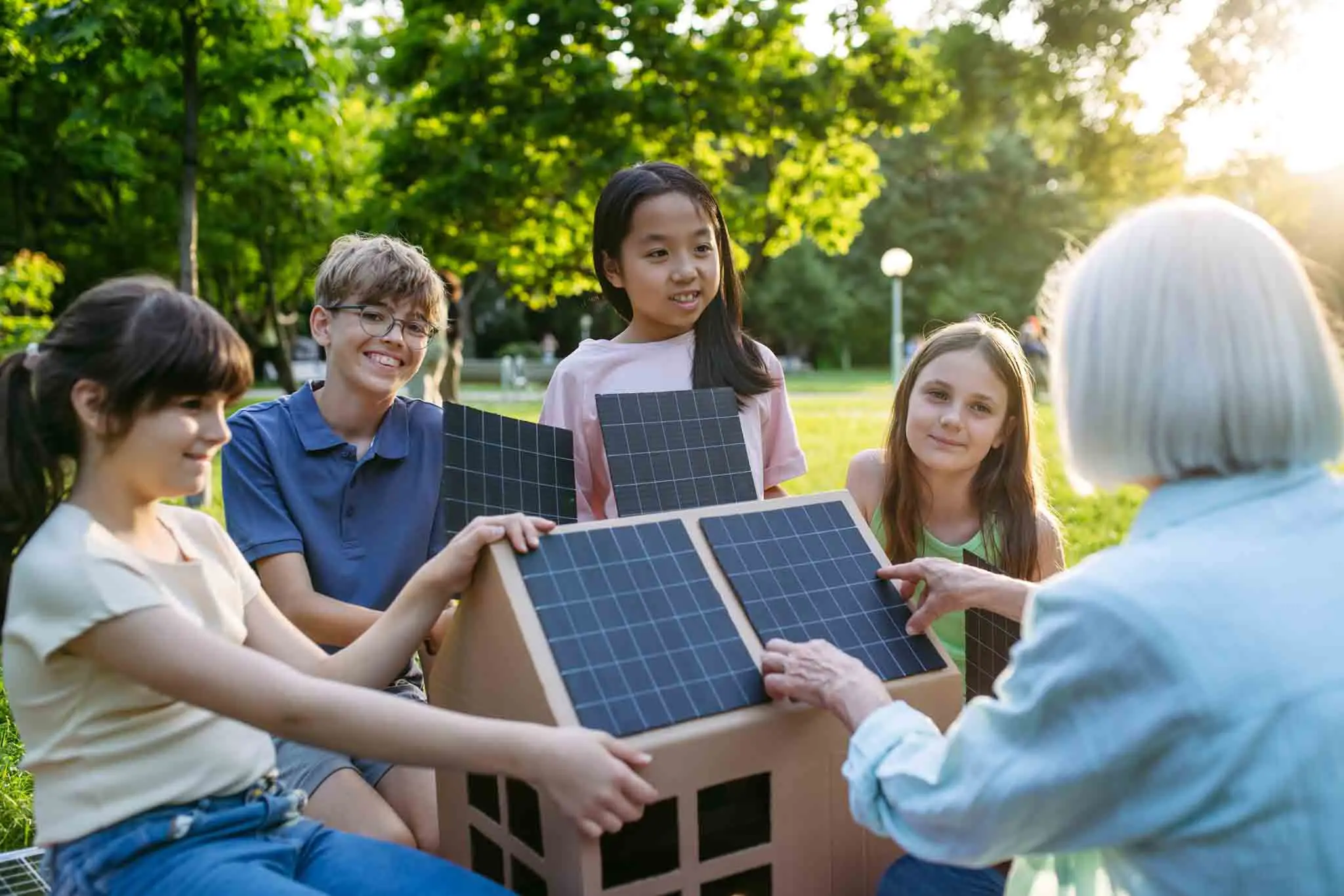 Children learning about renewable energy and solar panels during sustainable education class outdoors, using cardboard model of house wit solar panel on roof.
